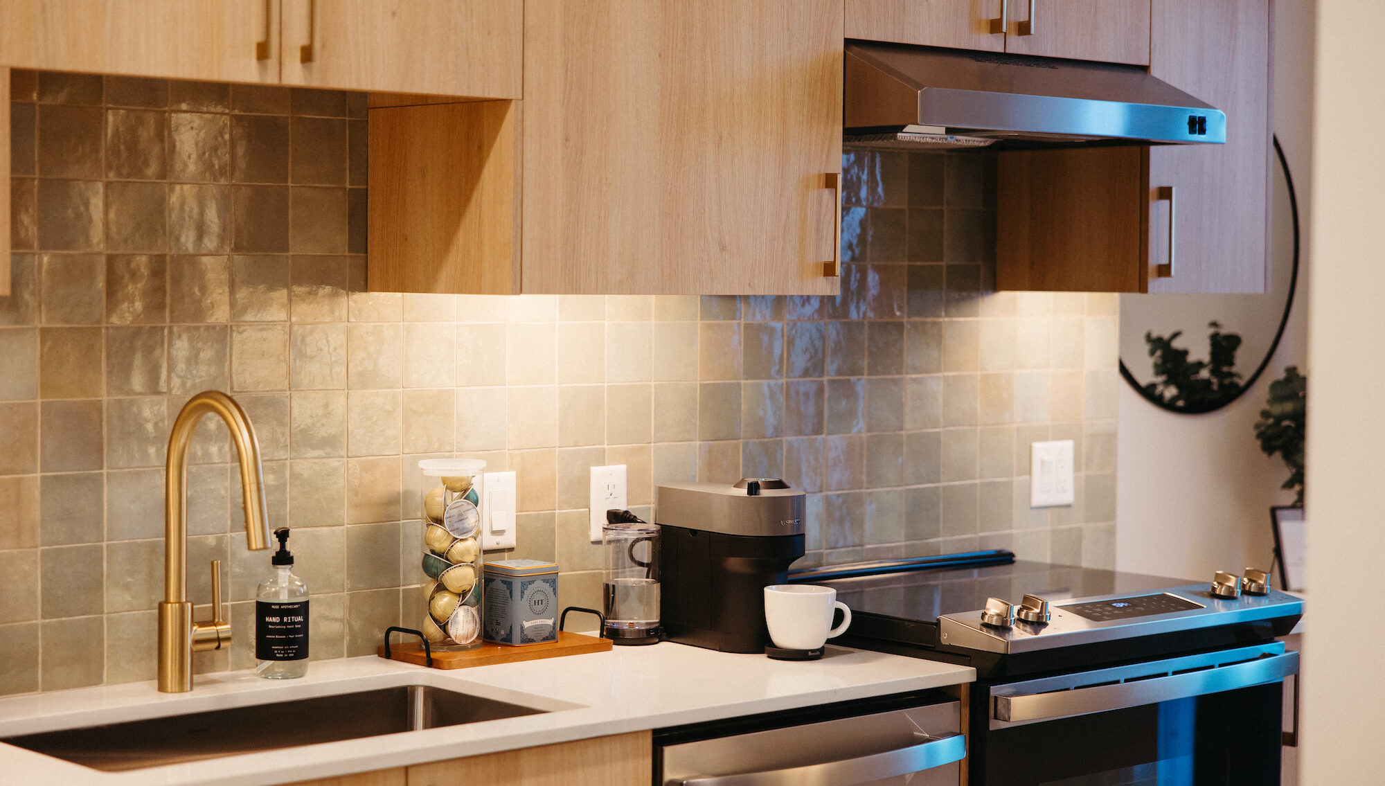 A modern kitchen in the guest suite features light wood cabinets, a tiled backsplash, gold faucet, and stainless steel appliances. A coffee maker, cup, soap dispenser, and jar of coffee pods rest on the white countertop.
